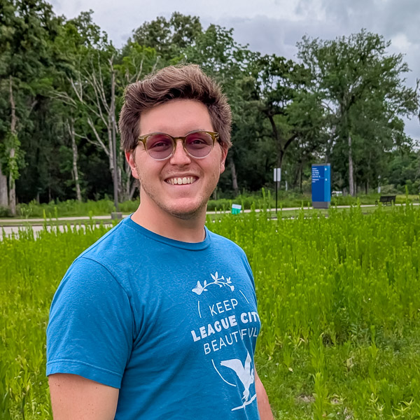 Smiling person in a blue t-shirt standing in a green field with trees and a cloudy sky in the background.