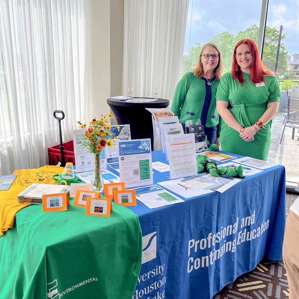 Two people stand behind an outreach table shared by EIH and UHCL’s PACE program, covered with brochures, flyers, and wildflowers.