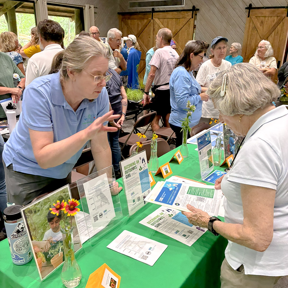 A person chats with a visitor at a table decorated with wildflowers and flyers, with other tables and people in the background.