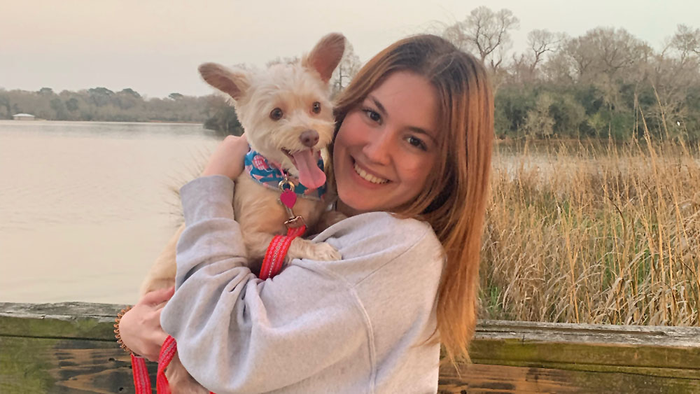 An individual, holding a dog, stand in front of a body of water, surrounded by tall grasses.