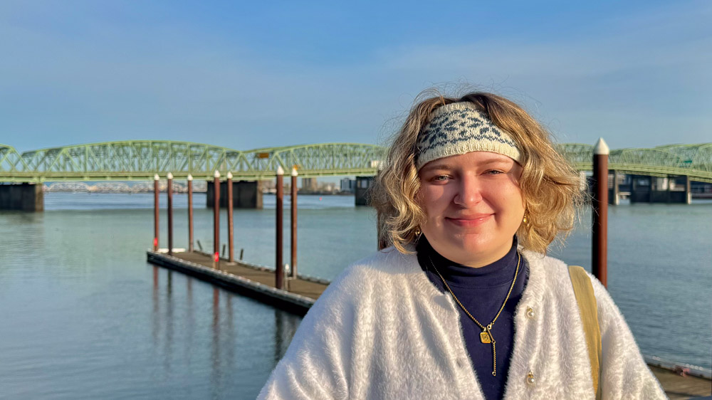 Person smiling near a riverfront dock with a green truss bridge in the background.