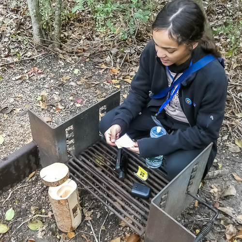 A young person squats near an inactive fire pit while holding a note found in a log-shaped geocache.