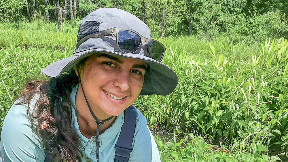 Person wearing a sun hat and field gear, smiling in a wetland area surrounded by dense green vegetation.