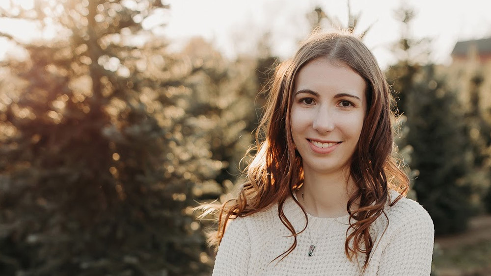 Person with long wavy hair wearing a light sweater, smiling outdoors with evergreen trees in the background.