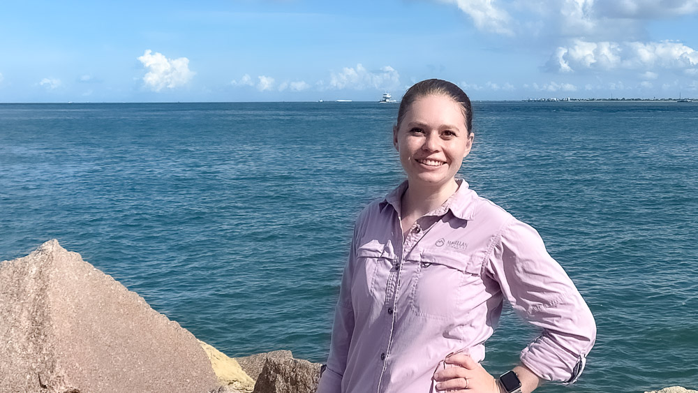 An individual standing on rocky shore with the ocean and a clear sky in the background.