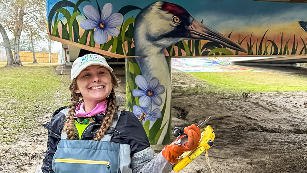 Chelsey is standing under a bridge with a mural of a whooping crane, blue-eyed grass, and blue metalmark in the background.