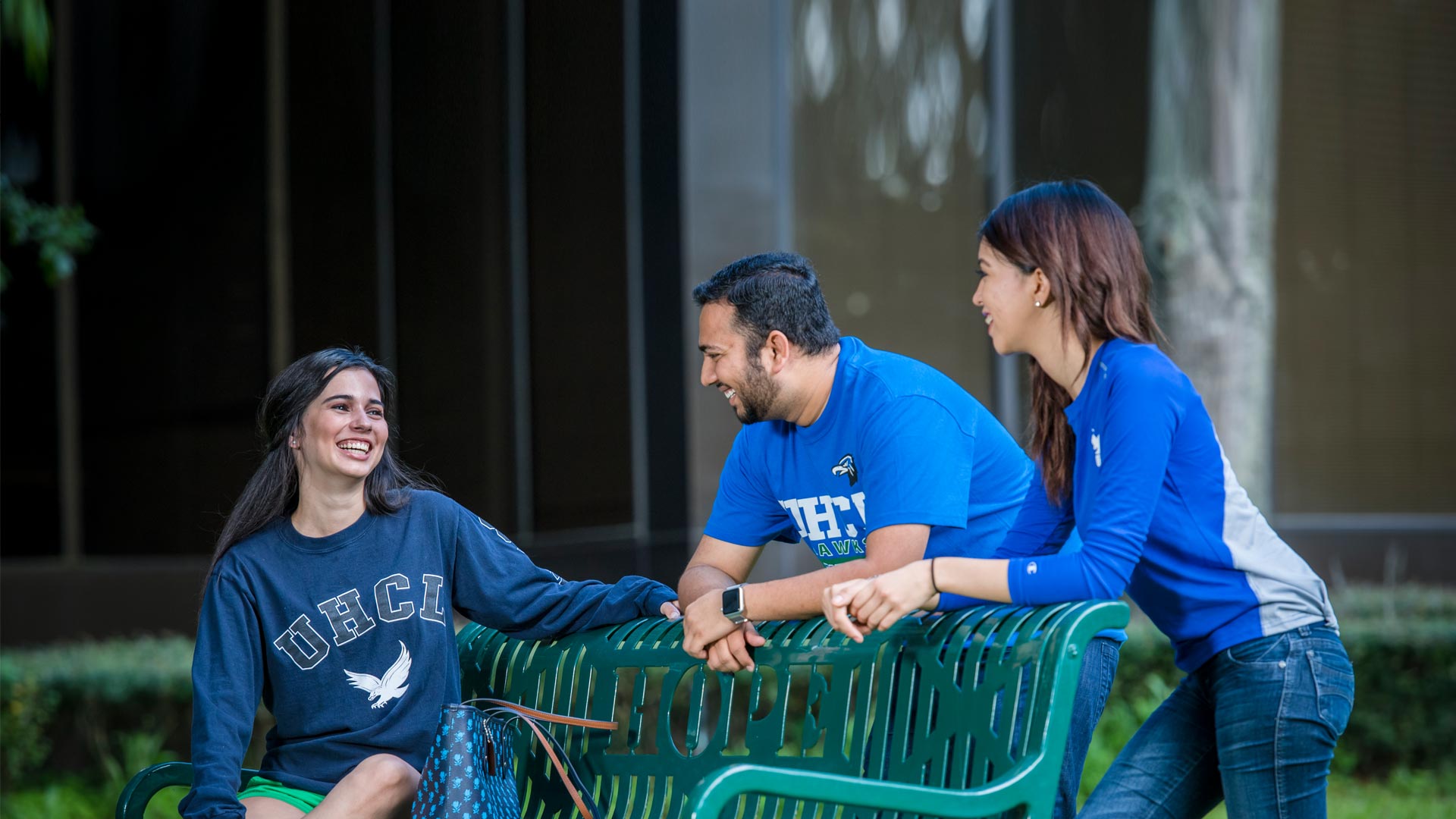 Three students engaged in a conversation while sitting on a bench outdoors, enjoying a sunny day at the University of Houston-Clear Lake.